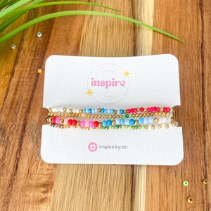 May include: A set of colorful beaded bracelets displayed on a white card. The bracelets feature a variety of colors, including red, pink, blue, and gold. The card has the word "inspire" printed on it, along with the Instagram handle "inspire.by.bri".