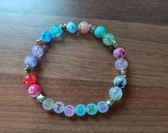 May include: A rainbow-colored beaded bracelet with gold stars and the word "STRONG" spelled out in colorful beads.