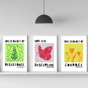May include: Three framed art prints with inspirational quotes. The first print features a green floral design with the words "JOY IS AN ACT OF RESISTANCE." The second print has a red butterfly with the words "HOPE IS A DISCIPLINE." The third print shows orange flowers with the words "LOVE IS AN ACT OF COURAGE."