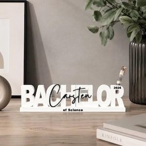 Könnte beinhalten: Ein weißes Schreibtischschild mit dem Wort BACHELOR in fetten Großbuchstaben, wobei der Name Carsten in schwarzer Schreibschrift in der Mitte darüber liegt. Der Text of Science steht darunter und das Jahr 2026 ist oben rechts aufgedruckt.