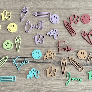 Könnte beinhalten: Eine Sammlung kleiner, lasergeschnittener Acrylformen, darunter Smileys, Bleistifte, Schultüten, Zahlen 123 und Buchstaben ABC, sowie verschiedene personalisierte Namen in Schreibschrift, die verstreut auf einer Holzoberfläche angeordnet sind.