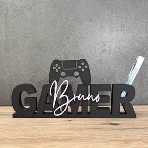 Könnte beinhalten: Schwarzer Schreibtisch-Organizer aus Holz mit dem ausgeschnittenen Wort "GAMER", einer Controller-Silhouette und dem Namen "Bruno" in Schreibschrift. Ein Stift befindet sich im "R" von "GAMER". Der Organizer steht auf einer Holzoberfläche.