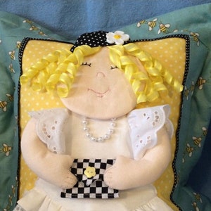 May include: A handmade doll with yellow curly hair, a white dress with lace trim, and a black and white checkered purse. The doll is wearing a black and white polka dot hat with a yellow flower. The doll is sitting on a blue and yellow pillow with a bee pattern.