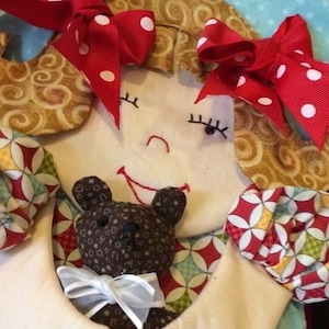 May include: A handmade doll with brown hair, wearing a red and white polka dot bow, a red and white patterned dress, and holding a brown teddy bear with a white bow.