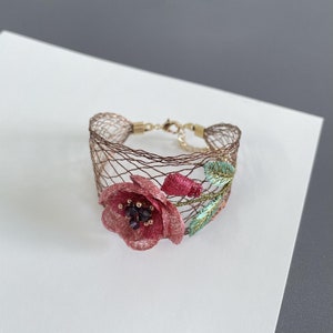 May include: A delicate bracelet made of a fine wire mesh with a pink embroidered flower and green leaves. The flower has a dark purple center.