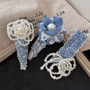 May include: Three blue denim hair clips with pearl accents. One clip features a blue flower with a pearl center and white lace trim. Another clip is adorned with a pearl strand and a silver charm. The third clip is a simple blue denim design with a pearl strand.