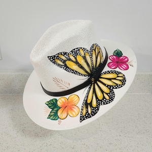 Può includere: Cappello di paglia bianco con fascia in pelle nera e un disegno di farfalla monarca dipinta. La farfalla ha ali gialle e nere con pois bianchi. Fiori rosa e arancioni con foglie verdi sono dipinti sul bordo.