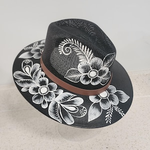 Op de afbeelding: Zwarte fedora hoed met een bruine band en wit bloemmotief. De hoed heeft een mesh kroon en een brede rand, met handgeschilderde witte bloemen en bladeren. Een stijlvol accessoire.