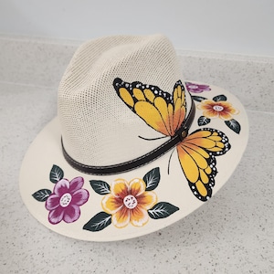 Peut inclure: Un chapeau de style fedora crème avec un bandeau en cuir marron. Le chapeau est décoré d'un grand papillon jaune et noir et de fleurs colorées. Le bord est orné de fleurs peintes dans des tons de violet, jaune et orange.