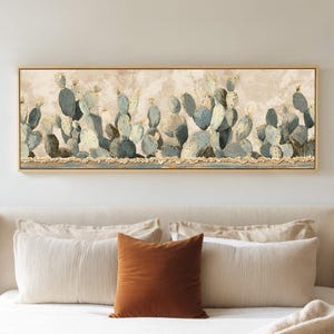 May include: A rectangular framed canvas art piece depicting a desert landscape with blue-green cacti. The artwork has a neutral color palette with a gold frame. The painting is displayed above a bed with white bedding and a brown pillow.