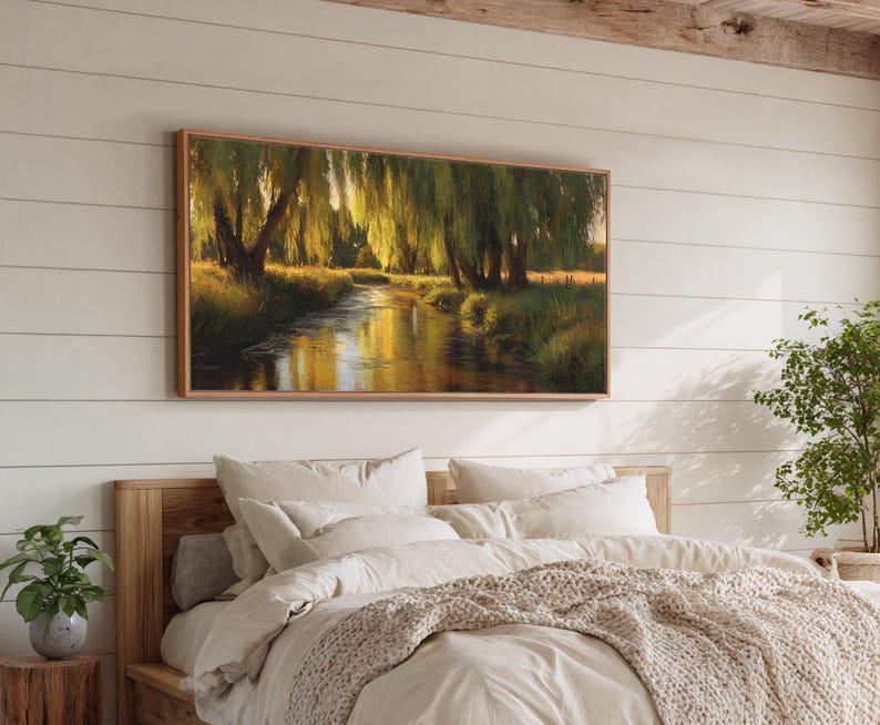 May include: A framed landscape painting depicts a serene river scene with weeping willow trees. The artwork features warm tones of gold, green, and brown, creating a tranquil and natural ambiance. The painting is displayed above a bed with neutral-colored bedding.