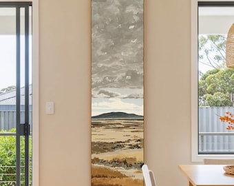 May include: Vertical landscape painting with a gold frame. The artwork depicts a coastal scene with a mountain in the distance, a sandy beach, and a cloudy sky. The color palette includes shades of brown, beige, gray, and blue.