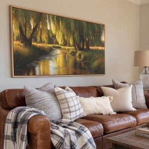 May include: A living room scene featuring a brown leather sofa adorned with decorative pillows and a plaid throw blanket. Above the sofa hangs a large rectangular framed painting depicting a serene landscape with a river and trees.
