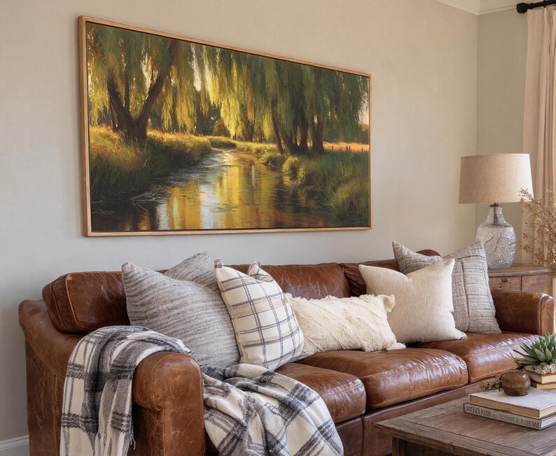 May include: A living room scene featuring a brown leather sofa adorned with decorative pillows and a plaid throw blanket. Above the sofa hangs a large rectangular framed painting depicting a serene landscape with a river and trees.