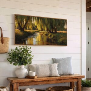 May include: A framed landscape painting depicts a serene river scene with weeping willow trees. The artwork hangs above a wooden bench with decorative pillows, a vase of greenery, and a woven tote bag. The wall is white with horizontal wood paneling.