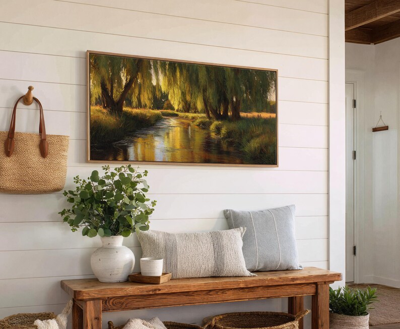 May include: A framed landscape painting depicts a serene river scene with weeping willow trees. The artwork hangs above a wooden bench with decorative pillows, a vase of greenery, and a woven tote bag. The wall is white with horizontal wood paneling.