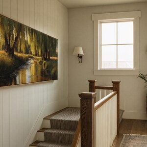 May include: A large rectangular framed painting depicting a serene landscape with a river flowing through a forest. The artwork is mounted on a white wall above a staircase with a neutral-toned runner. The scene is illuminated by natural light from a nearby window.