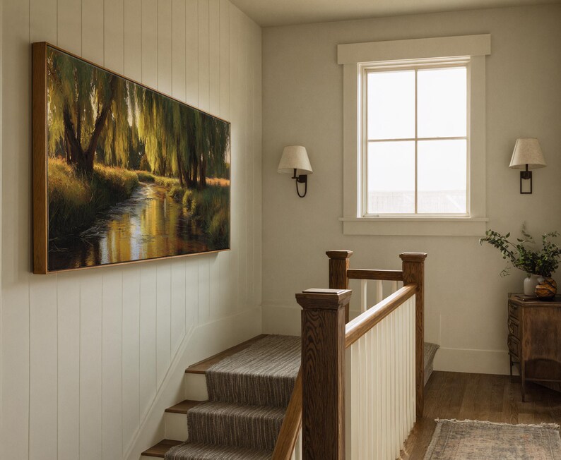 May include: A large rectangular framed painting depicting a serene landscape with a river flowing through a forest. The artwork is mounted on a white wall above a staircase with a neutral-toned runner. The scene is illuminated by natural light from a nearby window.