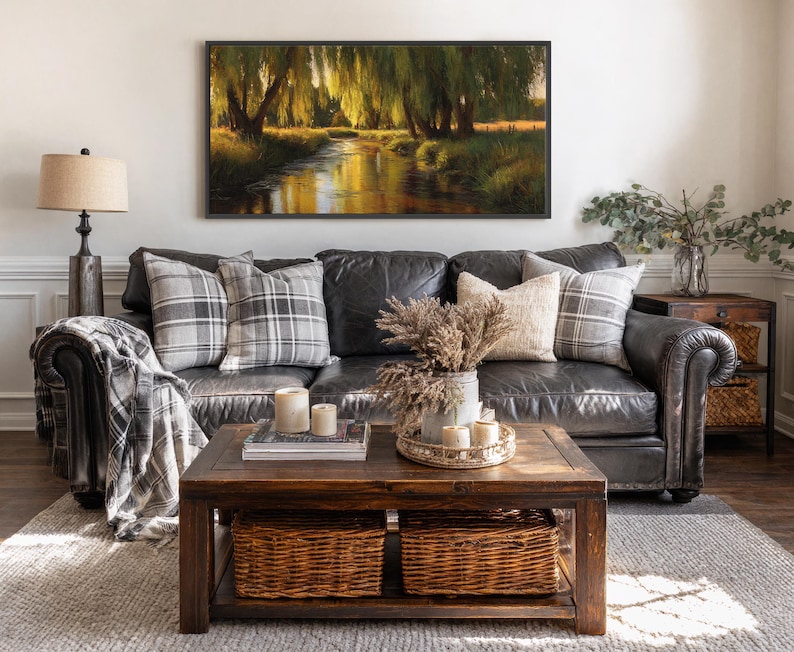 May include: A living room scene featuring a dark leather sofa with plaid pillows and a throw blanket. A wooden coffee table holds candles, books, and a tray with dried flowers. A framed landscape painting hangs above the sofa.