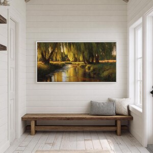 May include: A framed landscape painting depicts a serene river scene with weeping willow trees. The artwork, with a white frame, hangs above a wooden bench in a bright, white-walled hallway. A canvas tote bag hangs on the left.