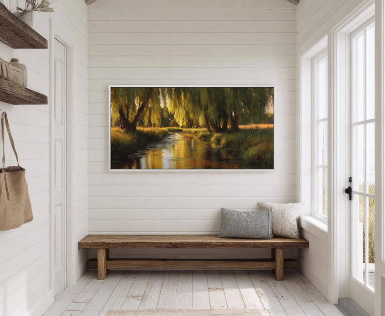 May include: A framed landscape painting depicts a serene river scene with weeping willow trees. The artwork, with a white frame, hangs above a wooden bench in a bright, white-walled hallway. A canvas tote bag hangs on the left.