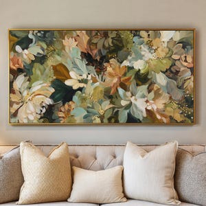 May include: A floral painting in a gold frame, featuring a mix of white, green, and peach-colored flowers and foliage. The artwork is displayed above a beige sofa with several decorative pillows, creating a warm and inviting atmosphere.