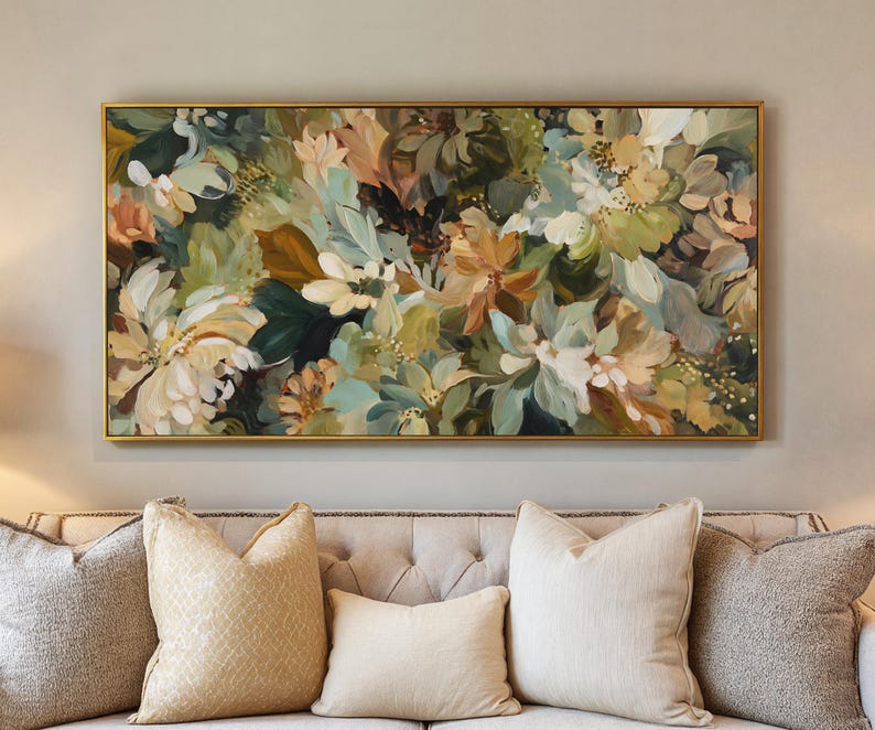 May include: A floral painting in a gold frame, featuring a mix of white, green, and peach-colored flowers and foliage. The artwork is displayed above a beige sofa with several decorative pillows, creating a warm and inviting atmosphere.