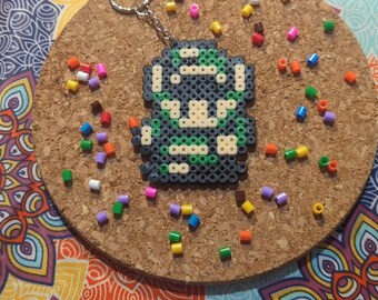 May include: A green, black, and white pixel art keychain of a video game character. The keychain is on a cork coaster with colorful beads scattered around it.