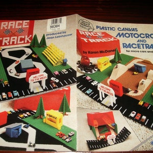 May include: A craft instruction booklet titled Plastic Canvas Motocross and Racetrack for micro cars and cycles by Karen McDaniel. The cover features photographs of completed plastic canvas race tracks, grandstands, and miniature vehicles.