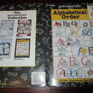 May include: A book cover featuring cross-stitch alphabet designs and other cross-stitch patterns. The cover includes the text "The good natured girls Collection" and "Alphabetical Order". The designs include letters, teddy bears, and floral patterns.