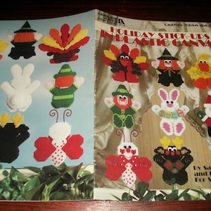 May include: A craft instruction booklet titled Holiday Stick-Ups in Plastic Canvas by Sandy Baggé and Honey Cain. The cover displays various plastic canvas figures including a turkey, witch, bunny, Santa, and butterfly in vibrant colors.