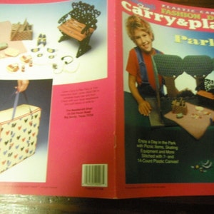 May include: A craft instruction booklet titled Carry & Play Fashion Doll Park featuring plastic canvas projects. The cover displays a park scene with dolls, a picnic blanket, trees, and a bench, alongside a carry case with a heart pattern and text.