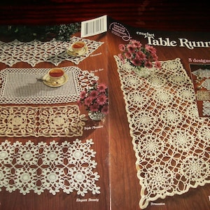 May include: A crochet pattern book titled Crochet Table Runners featuring 5 designs in thread. The cover displays various off-white lace table runners with intricate floral and geometric patterns, alongside photographs of finished projects.
