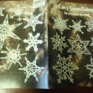 May include: A craft pattern booklet titled Lacy Snowflakes in crochet thread featuring various white, intricate, star-shaped snowflake designs on a dark background. The cover includes the text Lacy Snowflakes in crochet thread and produced by Rita Weiss.