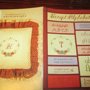 May include: A book titled "Script Alphabets" by Herriet True, featuring various embroidered alphabet styles. The book is open to a page with a pillow with the letter "H" embroidered on it, surrounded by a floral design.