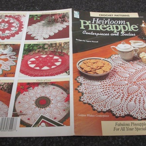 May include: A crochet pattern book titled "Heirloom Pineapple Centerpieces and Doilies" by Agnes Russell. The book features various crochet doily designs, including a white and red star-shaped doily and a white doily with a scalloped edge. A plate of crackers and a tea set are on the table.