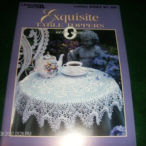 May include: A purple leaflet titled "Exquisite Table Toppers" by Leisure Arts. The cover features a white crocheted table topper on a table with a teapot, teacup, and statue. The leaflet's cover also includes the text "Souvenir Maid".