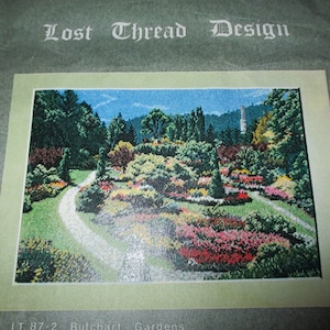 May include: A cross-stitch pattern featuring a detailed landscape of Butchart Gardens. The image shows a winding path through colorful flower beds, trees, and a tower against a blue sky. The text "Lost Thread Design" is at the top.