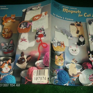 May include: A craft instruction booklet titled Plastic Canvas Magnets for Cat Lovers by Daria J. Fanton. The cover features numerous colorful cat-themed plastic canvas magnets, yarn balls, and metal cans. It includes a barcode and product number 3171.