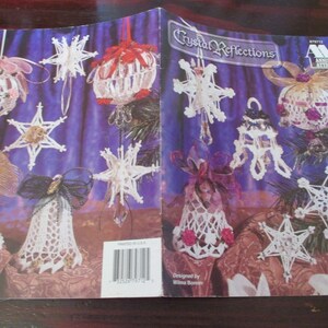 May include: A book cover titled "Crystal Reflections" featuring various handmade Christmas ornaments. The ornaments include crocheted snowflakes, bell shapes, and sphere designs, adorned with ribbons and beads. The color palette includes white, red, pink, and gold.