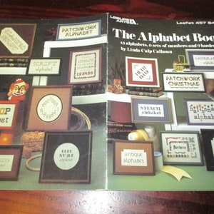 May include: A vintage craft book titled "The Alphabet Book" by Linda Culp Calhoun. The cover features framed cross-stitch samplers with various alphabet styles, including "Big Top," "Patchwork," and "Antique." The book includes 13 alphabets, 6 sets of numbers, and 9 borders.