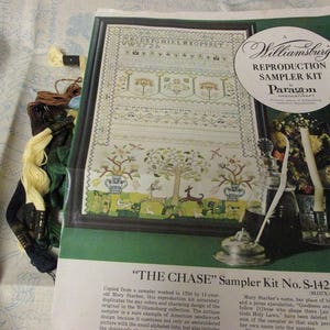 May include: A Williamsburg Reproduction Sampler Kit by Paragon Needlecraft titled The Chase, featuring a printed image of a cross-stitch sampler with alphabet, floral, and animal motifs, accompanied by several bundles of colored embroidery thread.