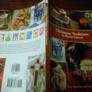 May include: A book titled "Christmas Traditions in Plastic Canvas" is open, displaying colorful needlecraft projects. The cover features a gingerbread house, ornaments, and a Santa Claus design. The back cover shows various Christmas-themed crafts.