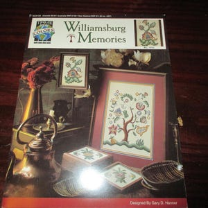 May include: A book cover with the title "Williamsburg Memories" and a floral design. The cover features framed cross-stitch patterns, a copper tea kettle, and a vase of flowers. The design is by Gary D. Hanner.