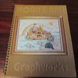 May include: A spiral-bound book with a gold cover featuring a framed image of Noah's Ark with a rainbow. The cover has vertical lines and the image shows animals on the ark. The book is on a dark brown wooden surface.