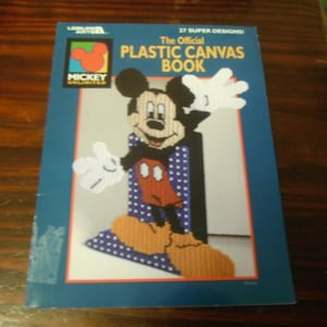 May include: A blue book titled "The Official Plastic Canvas Book" featuring a Mickey Mouse design. The cover shows Mickey Mouse in a stitched canvas design with white gloves, orange shoes, and a red and black outfit.
