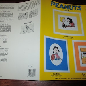 May include: A Peanuts Lucy counted cross stitch pattern booklet featuring three designs: Love Notes with a 14" x 12" frame size, and Trust Me and The Doctor Is In, both with 8" x 10" frame sizes. The yellow cover includes stitching instructions and diagrams.