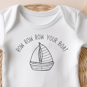May include: White baby onesie with the text "Row Row Row Your Boat" encircling a sailboat illustration. The onesie is short-sleeved and has snap closures at the bottom. The design is in black ink. The onesie is displayed on a woven basket.