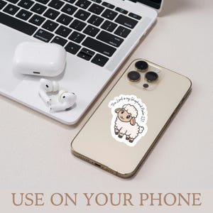 May include: A white sticker with a cartoon sheep and the text "The Lord is my Shepherd Psalm 23:1" is affixed to a gold smartphone. A laptop and white wireless earbuds are in the background. The phrase "USE ON YOUR PHONE" is at the bottom.