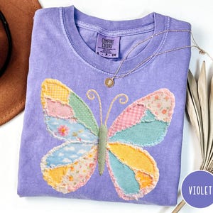 May include: A violet Comfort Colors t-shirt featuring a patchwork butterfly design. The butterfly has wings in various colors and patterns, including yellow, pink, blue, and floral prints. A gold necklace with a pendant rests on the shirt. The shirt is labeled "Violet".